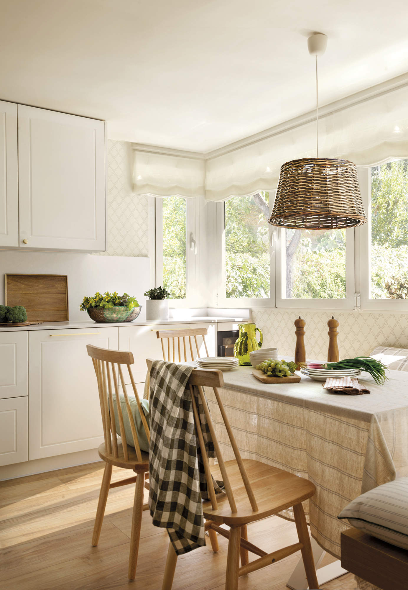Cocina blanca con office y suelo de madera, ventanas con estor blanco, mesa con banco y silla de madera, lámpara de techo de fibra, mantel de lino a rayas